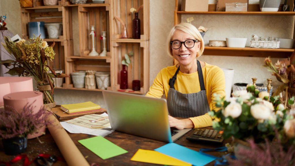 A woman providing website design for small businesses, sitting at a desk with flowers and a laptop.