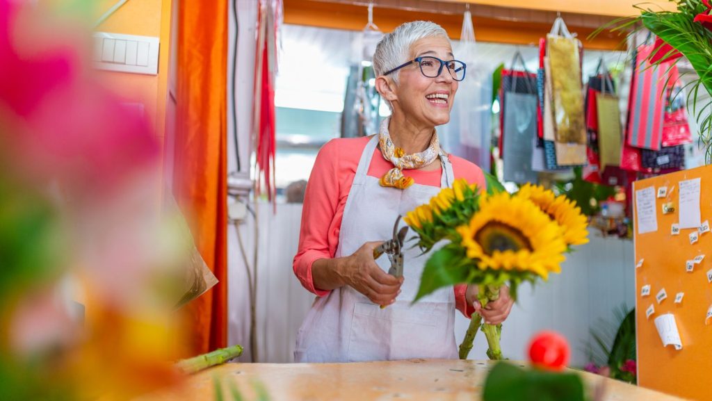 An older woman is holding sunflowers in a small flower shop working with a small business web designer.