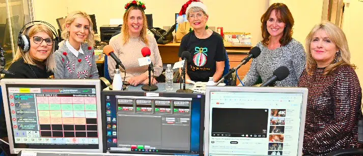 Six women sitting at a Brooklands Radio station desk with microphones and computer monitors in front of them, some wearing festive attire including santa hats, appear to be hosting or participating in a live broadcast.