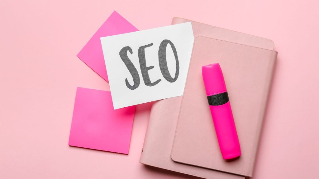 A pink highlighter, pink note cards, and a paper labeled "SEO" are placed on a pink folder against a pink background.