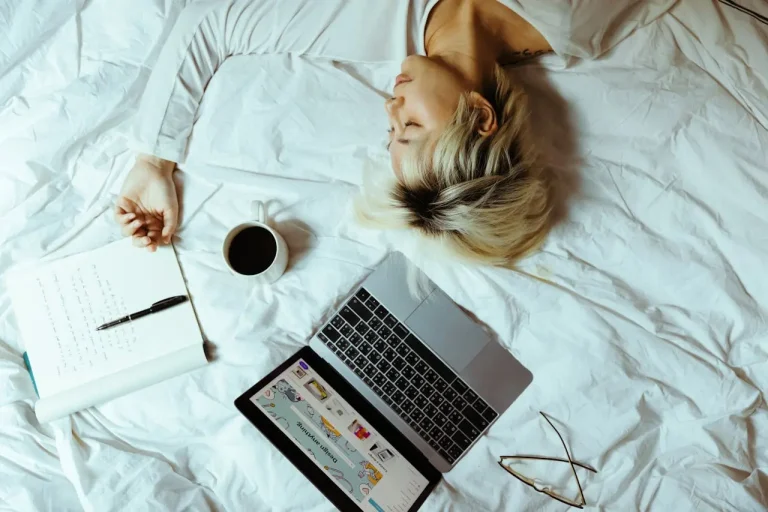 Person lying on a bed beside an open laptop displaying what a website is not, a notebook with writing, a pen, a pair of glasses, and a cup of coffee.