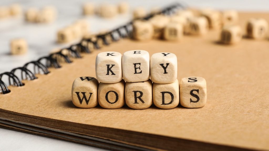 Wooden dice with letters arranged to form the words "KEYS" and "WORDS" are placed on a notebook with spiral binding, subtly illustrating the concept of keyword stuffing.