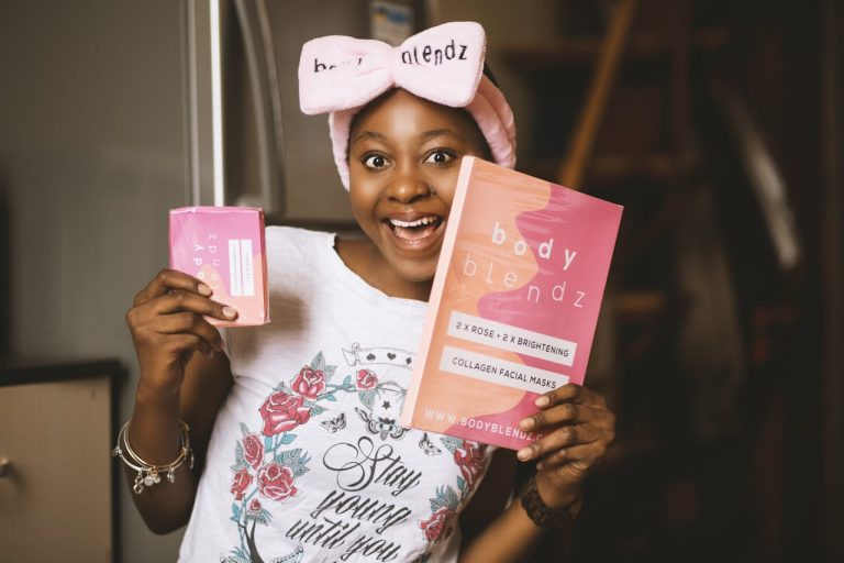 A person wearing a pink headband beams with emotion, holding a box and a packet of Body Blendz collagen facial masks.