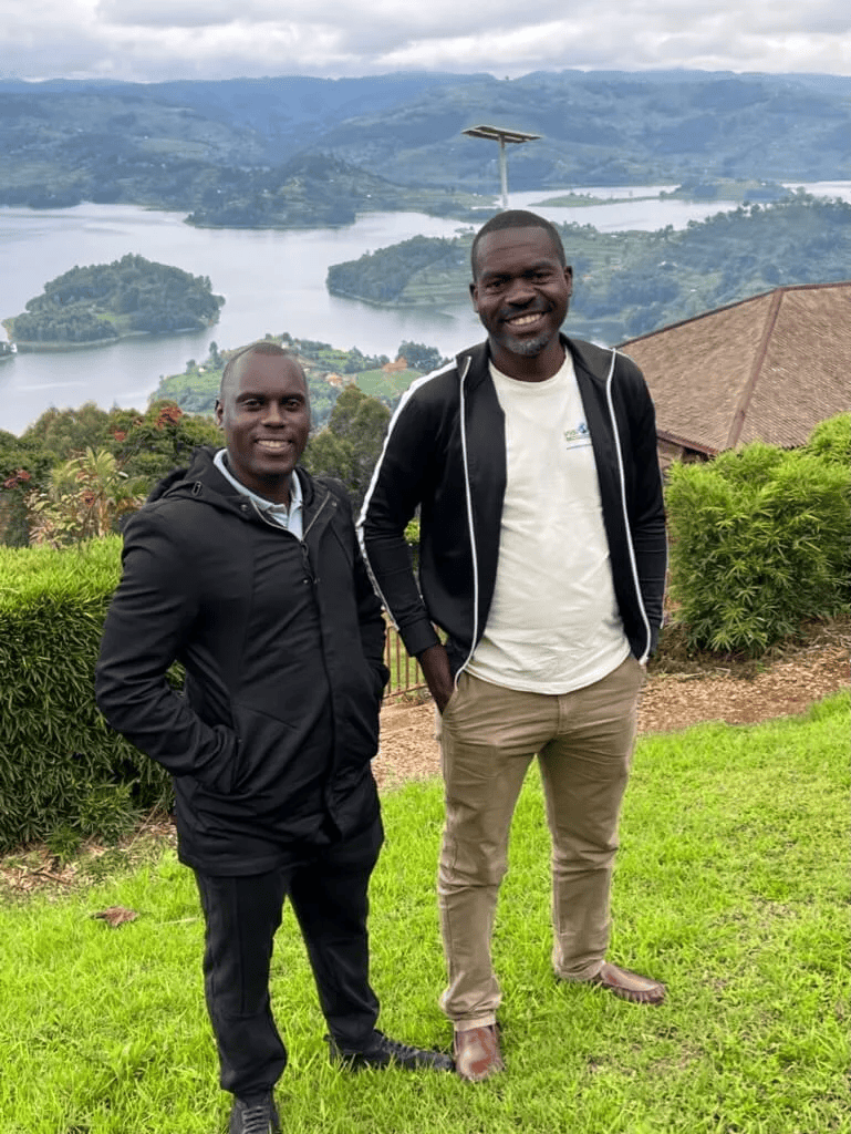 Two men stand outdoors on a grassy area with a scenic view of a lake and hills in the background. They are smiling and wearing casual clothing with jackets.