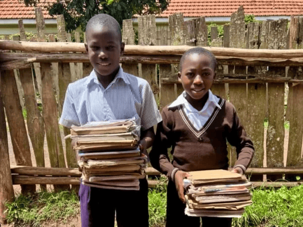 Two children standing outdoors, each holding a stack of books. A wooden fence and building with a red roof are in the background.