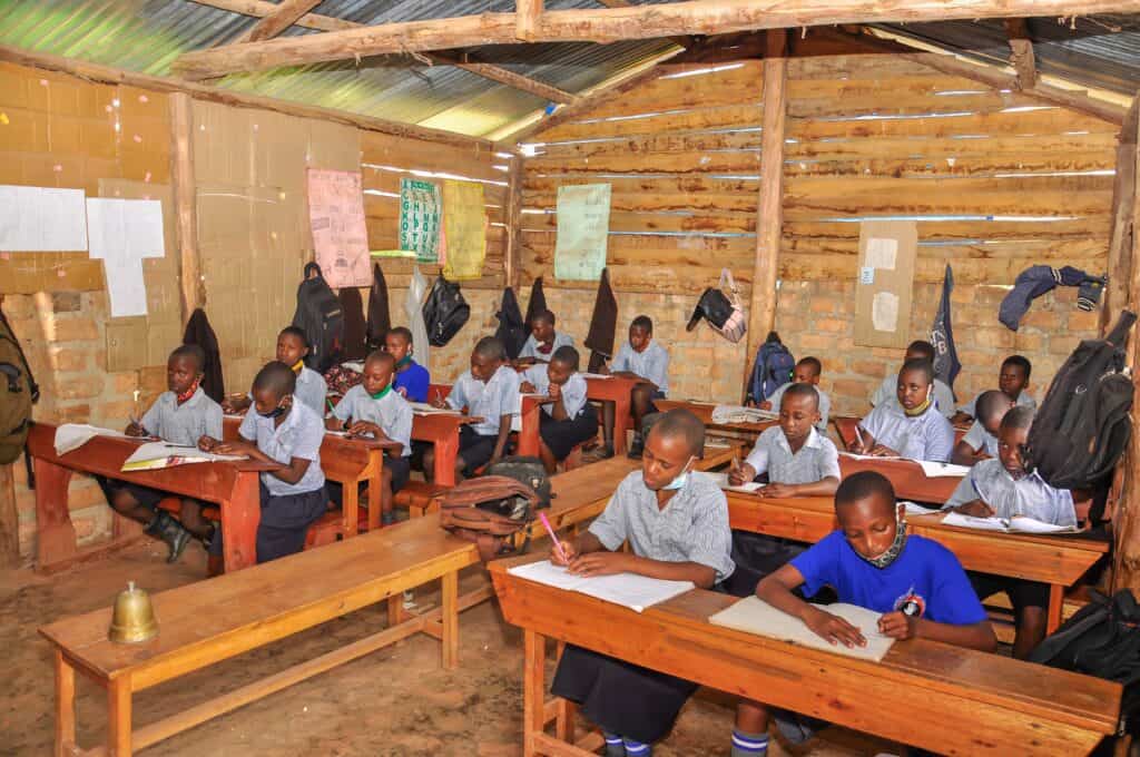 Students in uniforms sit at wooden desks and write in notebooks inside a rustic classroom with wooden walls and a metal roof.