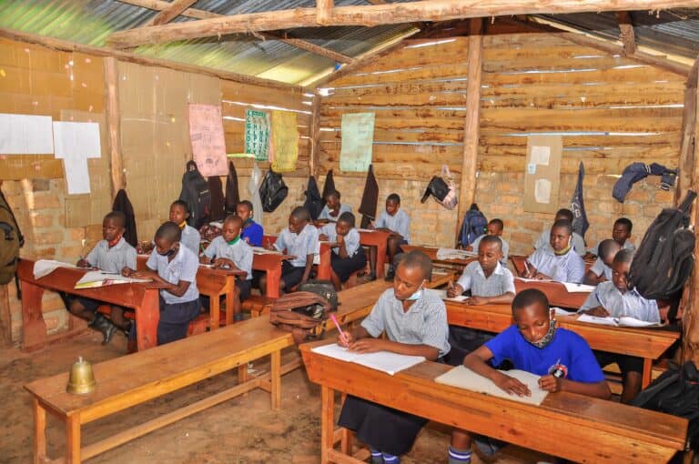 Students in uniforms sit at wooden desks and write in notebooks inside a rustic classroom with wooden walls and a metal roof.