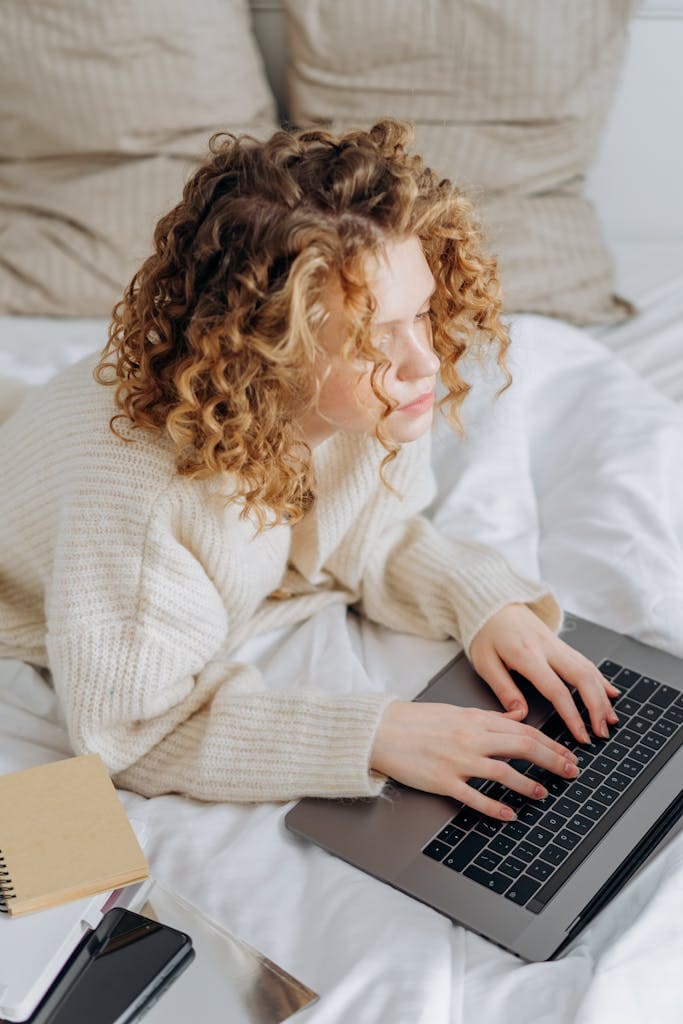 A young woman with curly hair typing on a laptop in bed, epitomizing remote work vibes. Website VIP Day