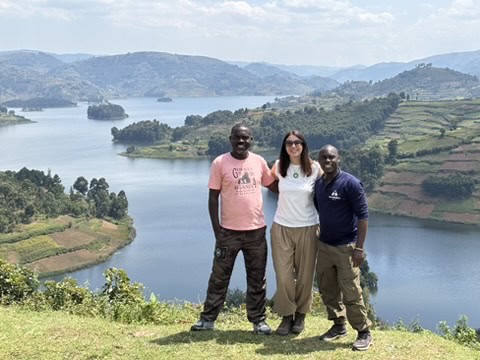 Three people stand smiling on a grassy hilltop overlooking a large lake and terraced hills under a partly cloudy sky, enjoying the scenic beauty with Amatsiko Tours.