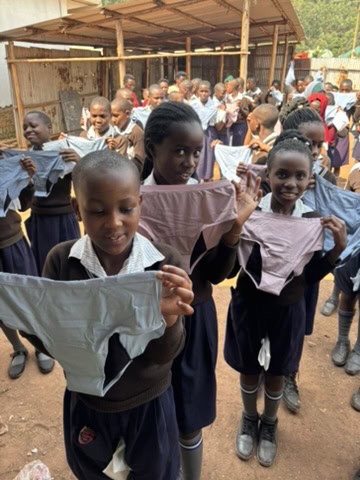 A group of schoolgirls in uniform stand outdoors during an Amatsiko Tours event, each holding up a new pair of underwear and smiling at the camera.