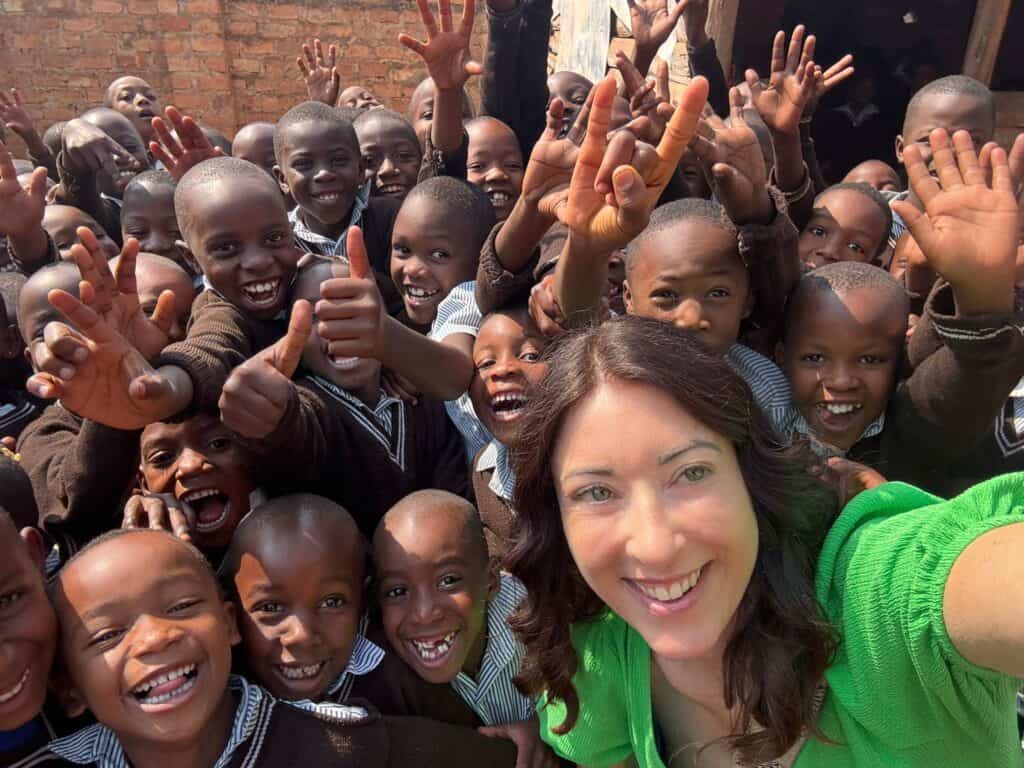 A woman in a green shirt takes a selfie with a large group of smiling children in school uniforms, all looking at the camera and waving during an Amatsiko Tours visit.