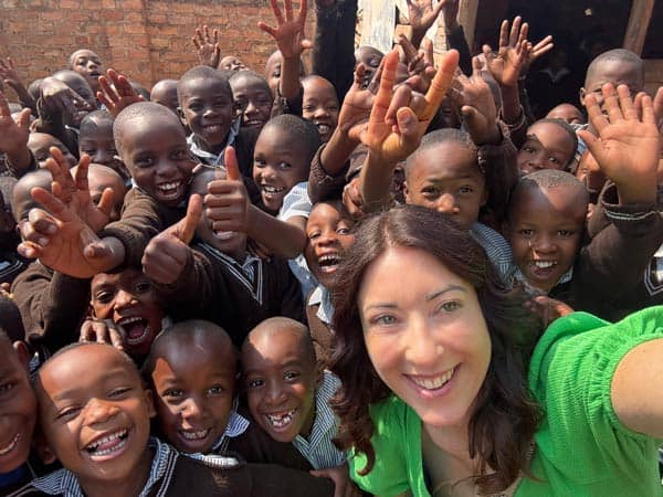 A woman in a green shirt takes a selfie with a large group of smiling children waving and giving thumbs up, capturing the joyful spirit of Amatsiko Tours against a brick wall backdrop.