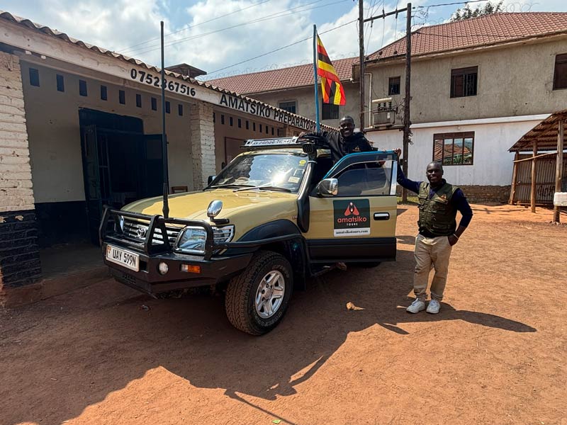 Two people stand by an Amatsiko Tours safari vehicle in front of a building labeled "Amatsiko Preparatory School." One person leans out of the driver’s window, with a flag attached to the vehicle.