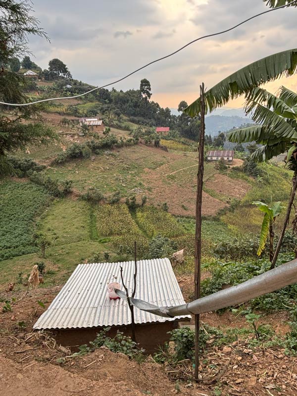 A corrugated metal roof is in the foreground, with sloped farmland, trees, and scattered houses stretching across a hilly rural landscape under a cloudy sky—an authentic scene you might discover on an Amatsiko Tours adventure.