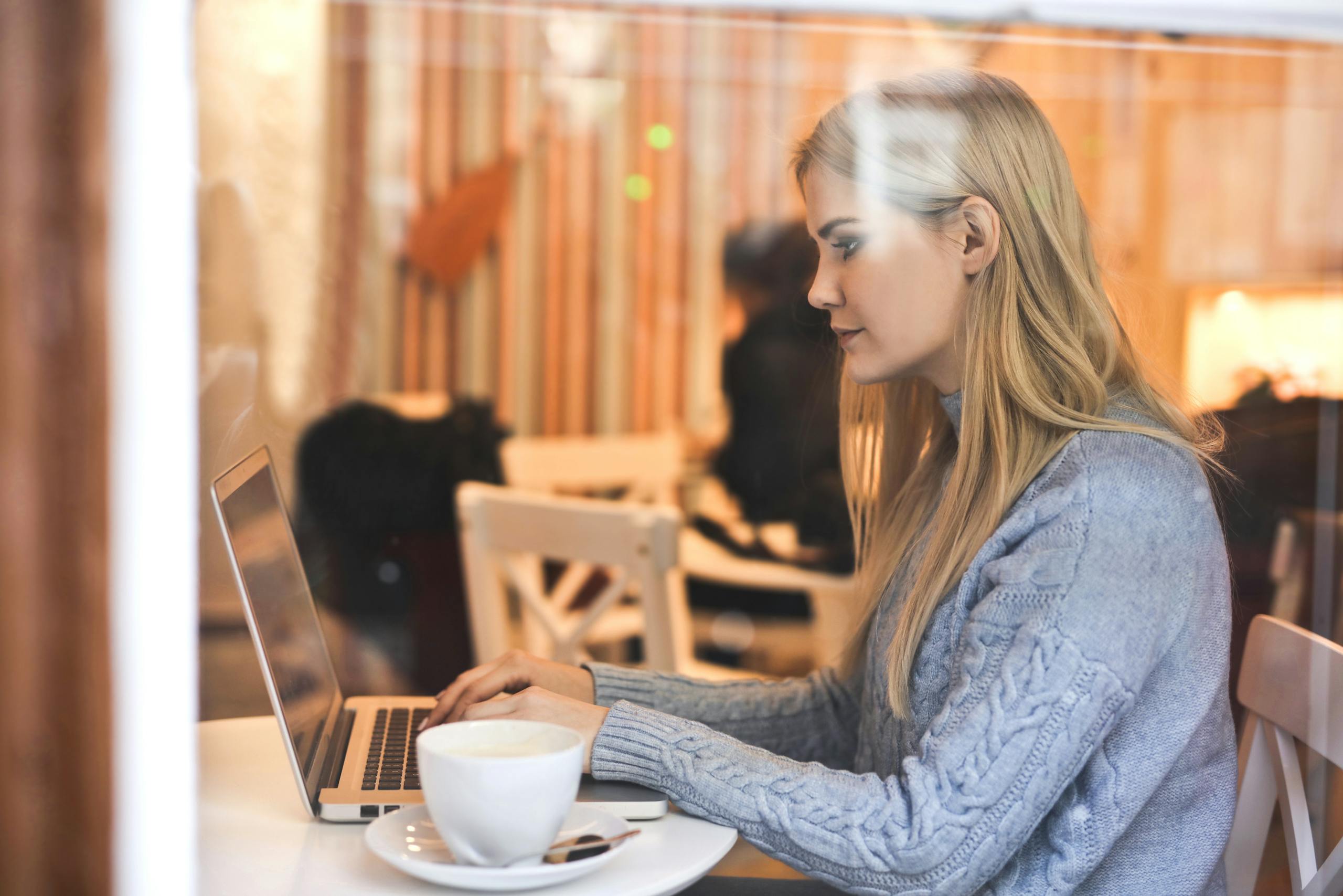 Blonde woman in a blue sweater using a laptop in a cozy café setting.