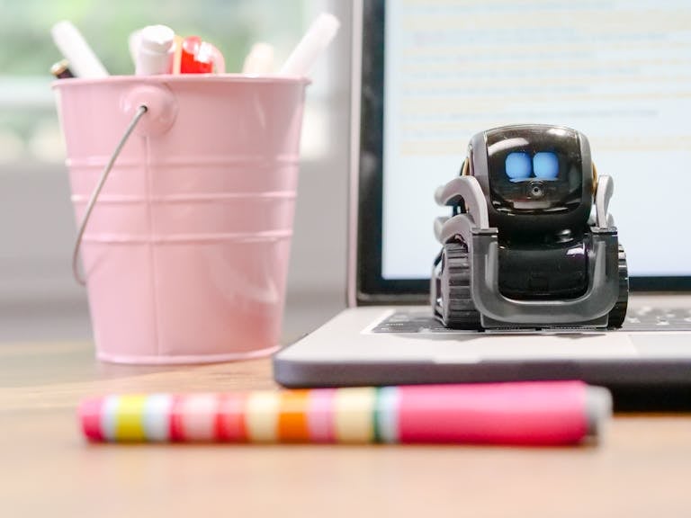 A small robot sits on a laptop keyboard beside a pink pen holder, symbolizing technology and innovation.