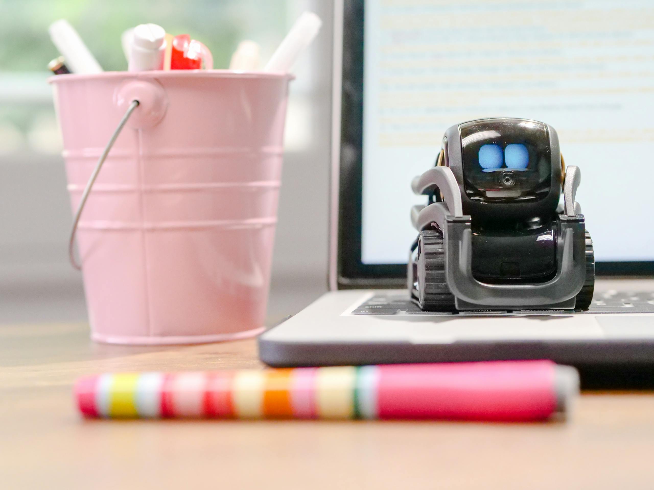 A small robot sits on a laptop keyboard beside a pink pen holder, symbolizing technology and innovation.